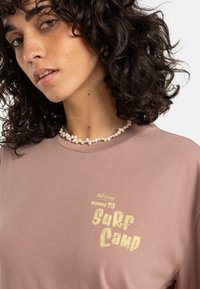 lichtroze katoenen t-shirt met 'Surf Camp' graphic in goud, ronde halslijn en korte mouwen, gedragen met een parelketting.