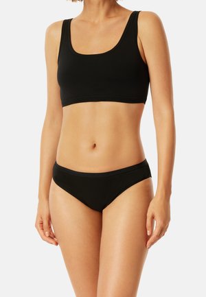 Frau trägt einen schwarzen Sport-BH mit Rundhalsausschnitt und passende schwarze Bikini-Unterwäsche, sichtbar vom Nacken bis zu den oberen Oberschenkeln.