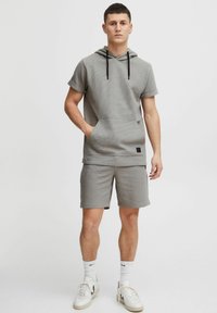Sudadera gris de manga corta con un bolsillo frontal y cordón negro. Combinada con pantalones cortos a juego, ambos con un patrón texturizado.