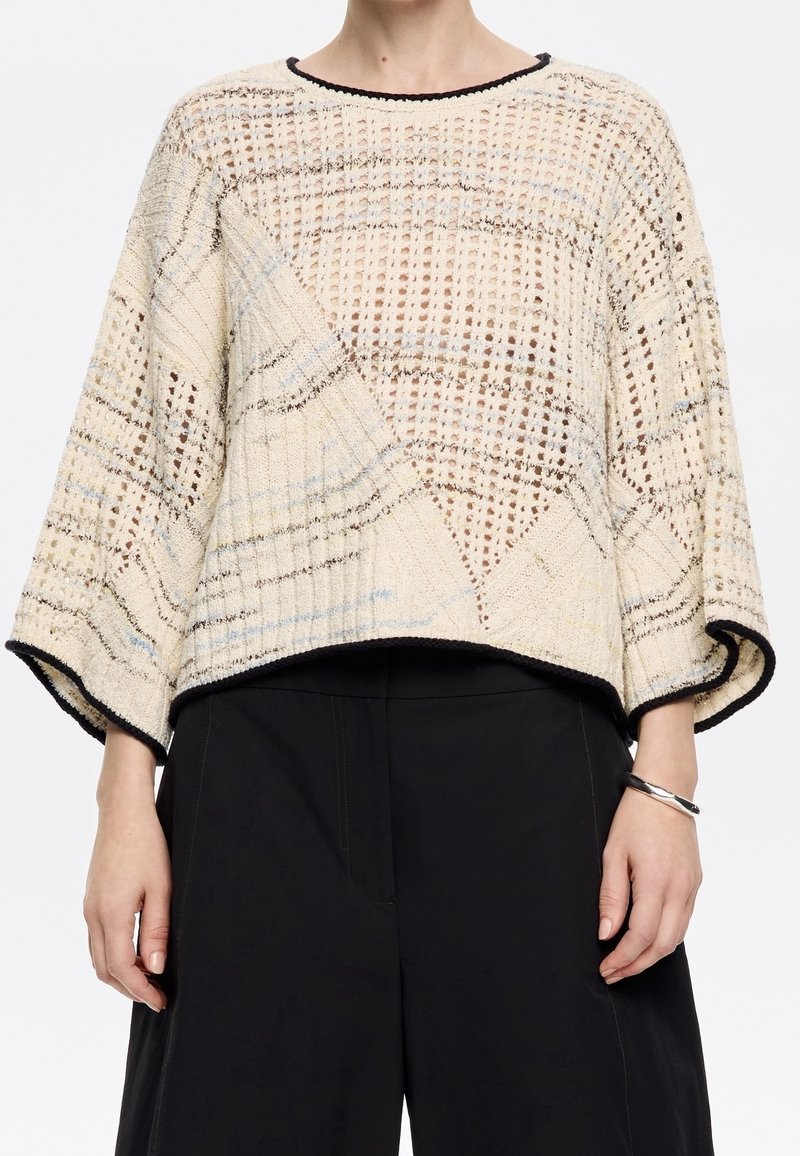 Femme portant un pull en maille beige ample avec une bordure noire et des manches larges, associé à un pantalon noir et un bracelet argenté au poignet droit.