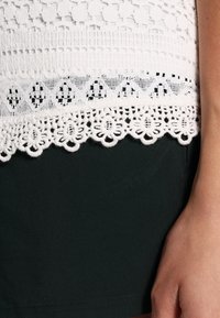 Top de ganchillo blanco con intrincados detalles de encaje en el dobladillo, con un patrón texturizado y un fondo verde oscuro sólido.