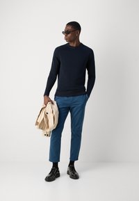 Marineblaue strukturiert gestrickte Pullover, petrolblaue Slim-Fit-Hosen, schwarze Schnürschuhe, hält eine beigefarbene Jacke. Minimalistisches Design mit kontrastierenden Farben.