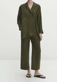 Giacca avvolgente in lino verde oliva con maniche lunghe e cintura, abbinata a pantaloni larghi coordinati. Indossata con infradito nere.