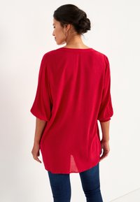 Rode blouse met een gekreukte textuur, losse pasvorm en driekwart mouwen. Heeft een ronde halslijn en een langere achterzoom.