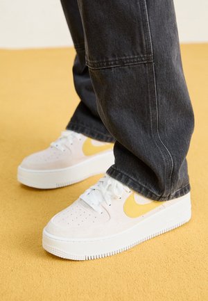 White sneakers with yellow Nike swoosh worn with dark gray jeans on a yellow carpeted floor.