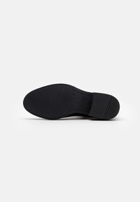 Suela de zapato de goma negra con superficie texturizada, que presenta patrones acanalados para un mejor agarre. Bordes redondeados y área del tacón reforzada.