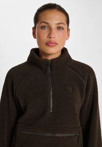 Brun fleece-pullover med hög krage, kvart dragkedja fram och en framficka med dragkedja. Har ett texturerat och mysigt tyg.