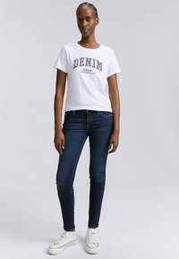 Weißes Baumwoll-T-Shirt mit dem Text "DENIM CREW" in Marineblau, kombiniert mit dunkelblauen Jeans und weißen Sneakern. Das Modell steht aufrecht vor einem grauen Hintergrund.