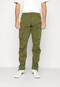 Mies, joka wear olive green cargo pants useilla taskuilla ja valkoisilla tennareilla yksinkertaista valkoista taustaa vasten.