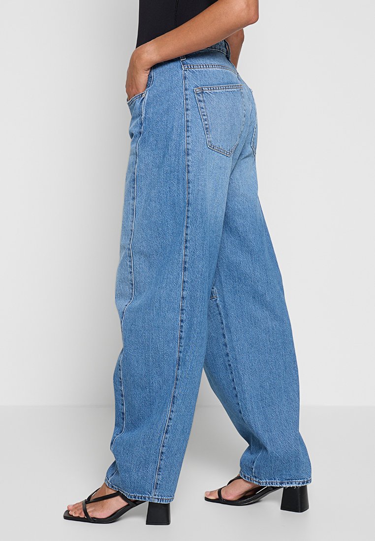 Jasnoniebieskie dżinsowe spodnie o szerokich nogawkach i wysokim stanie, z tylnymi kieszeniami i kontrastowym przeszyciem, noszone z czarnymi sandałami na paski.