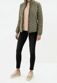 Frau in einer olivgrünen gesteppte Jacke über einem Pfirsichpullover, schwarzen Skinny Jeans und beigen Sneakers mit weißen Sohlen vor einem weißen Hintergrund.