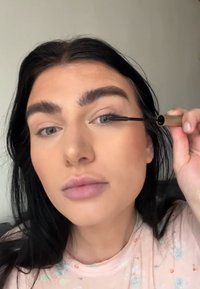 Du mascara noir appliqué avec un tube doré, mettant en valeur des cils longs et foncés. La personne a la peau claire, des lèvres pâles et des cheveux foncés.