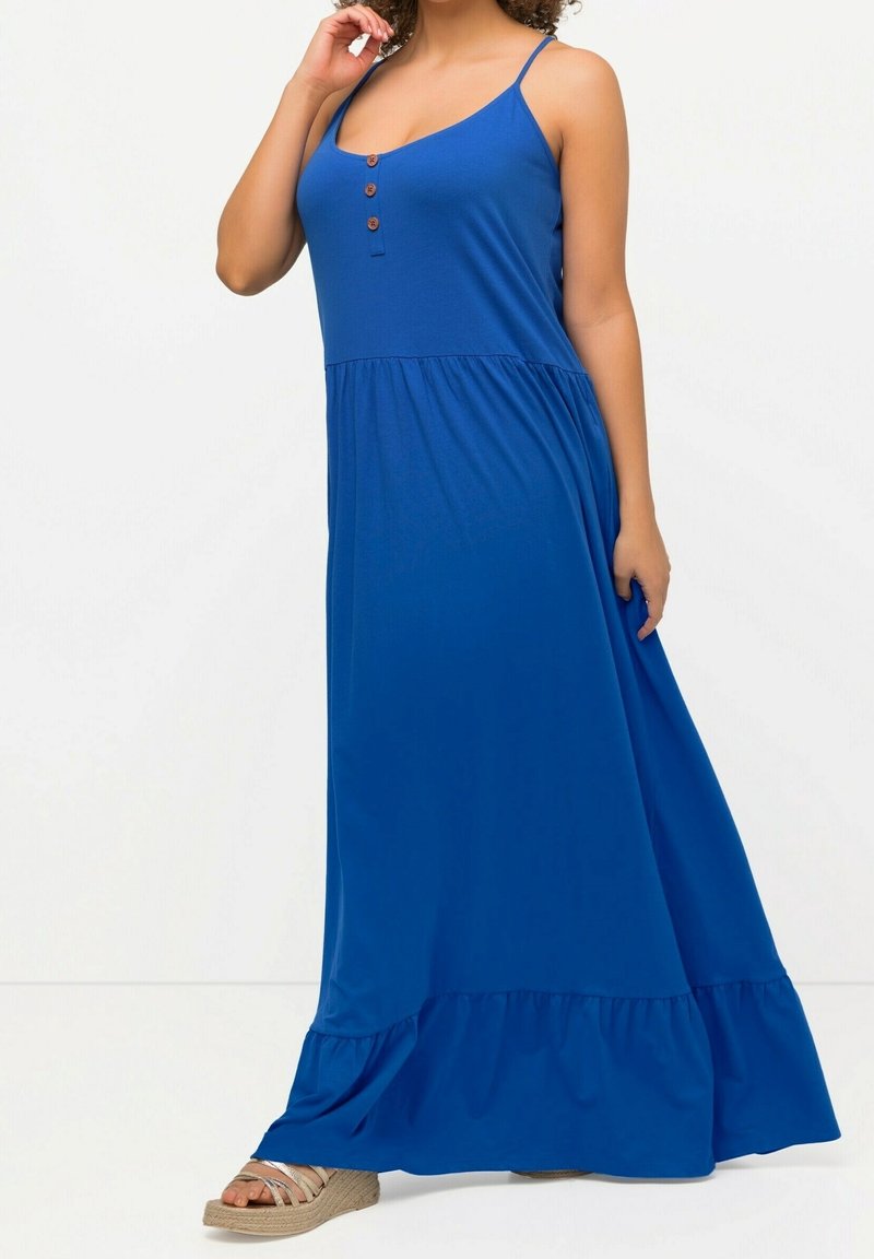 Femme portant une robe maxi bleu royal sans manches avec fines bretelles, boutonnage sur le devant, ourlet à volants et sandales compensées beige.