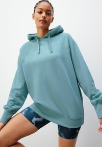 Haut de sweat à capuche bleu clair avec cordon de serrage, poignets et ourlet côtelés. Le tissu semble doux. Porté avec un short foncé à motifs.
