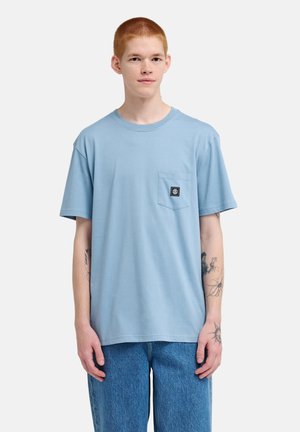 Jonge persoon met kort rood haar draagt een lichtblauw T-shirt met zakken en blauwe spijkerbroek, met tatoeages op beide onderarmen zichtbaar.