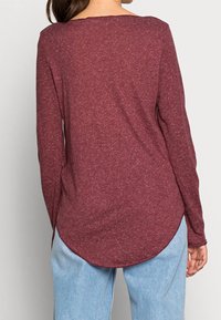 Långärmad topp i mörk borgonya, strukturerad tyg med rundad nederkant. Matchad med ljusblå jeans. Sömmar är tydliga längs ärmarna.