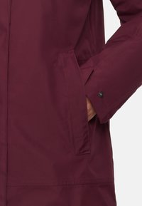 Ręka w kieszeni długiego, ciemnofioletowego kurtki z długim rękawem, zapinanej na nap na mankiecie, na białym tle.