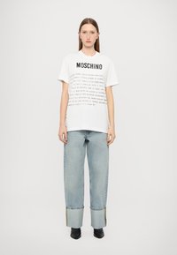 T-shirt branca com o texto preto "MOSCHINO" à frente, combinada com jeans de denim azul claro com um corte largo e bainhas desfiadas.
