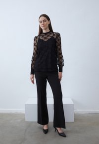 Black lace top with long sheer sleeves, a round neckline, paired with black tailored pants and black pointed heels. Smooth textured background.