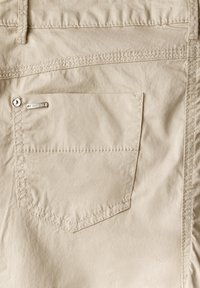 Tascone posteriore in tessuto beige con un'accensione del logo in metallo, caratterizzato da bordi cuciti e forma rettangolare. Texture liscia e design semplice.