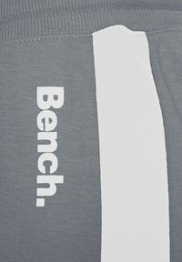 Graue Stoff-Jogginghosen mit einem weißen Seitenstreifen und dem Wort "Bench." in fettem weißen Text auf dem linken Bein. Weicher Stoff, lässiges Design.