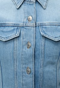 Φωτεινό γαλάζιο τζιν μπουφάν με μεταλλικά κουμπιά και δύο τσέπες καπακιού στο στήθος, εμφανείς ραφές.