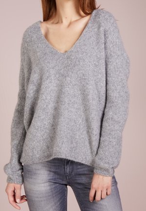Vrouw die een loszittende, lichtgrijze V-hals trui met lange mouwen draagt en vervaagde grijze spijkerbroek, tegen een neutrale achtergrond.