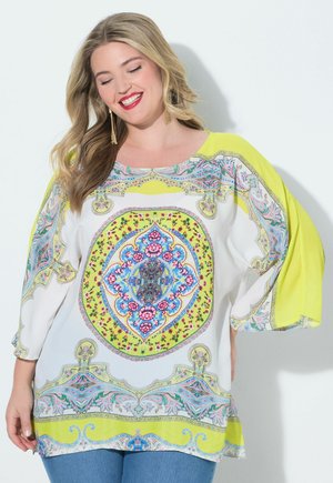 Lächelnde Frau mit langen Haaren, die goldene Ohrringe und eine weiße Tunika mit gelben und blauen Mustern sowie floralen und geometrischen Designs trägt.