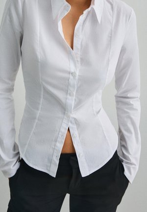 Dopasowana biała koszula zapinana na guziki, lekko rozpięta u góry i na dole, noszona z czarnymi spodniami, osoba stoi z rękami w kieszeniach.