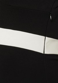 Tecido preto com uma proeminente faixa horizontal branca. Apresenta um detalhe de bolso com fecho seguro, exibindo uma textura suave e um design em malha.