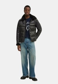 Giacca a vento nera con colletto in pelliccia sintetica, abbinata a una maglietta blu navy, jeans dritti azzurri chiari e stivali neri su uno sfondo neutro.