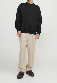 Osoba oblečená v černém oversized svetru, béžových širokých kalhotách a tmavě hnědých slip-on mokasínech stojí na bílém pozadí.