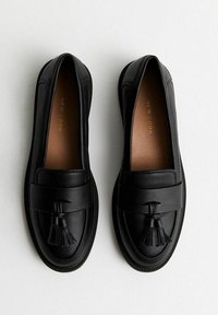 Mocassini in pelle nera con suola piatta, caratterizzati da una nappa decorativa. Texture liscia, punta arrotondata e dettagli di cucitura sottili.