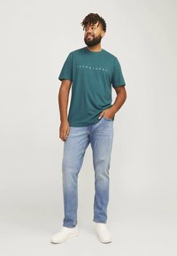 Tumši zaļa kokvilnas t-krekls ar apaļu kaklu un baltu logotipa tekstu, kopā ar gaiši zilām džinsa biksēm un baltām sporta kurpēm. Ikdienišķs piegriezums.