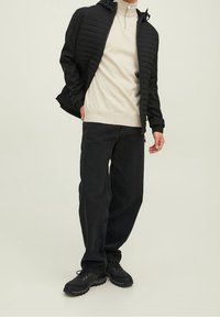 Veste matelassée noire avec capuche, portée sur un pull col montant beige clair, associée à un jean noir ample et des baskets noires.