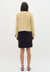 Keltainen ja valkoinen houndstooth-kuvioitu takki, jossa on pitkät hihat, lyhyt malli, yhdistetty mustaan, polvenpituiseen hameeseen, jossa on sivutaskut.