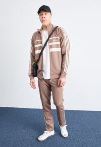 Brown zip-up track jacket with cream stripes, paired with matching brown track pants. White t-shirt underneath, black cap, and white shoes.