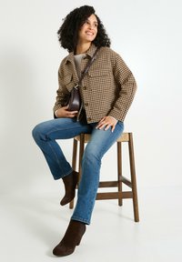 Veste à motif pied-de-poule marron, jeans bleus et bottes chelsea marron. Pose sur un tabouret en bois, avec un petit sac crossbody marron.