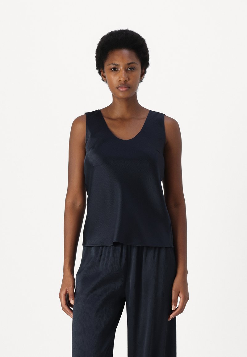 Marineblaue ärmellose Bluse aus glattem, glänzendem Stoff mit rundem Ausschnitt und lockerer Passform, kombiniert mit dunklen, gefalteten Hosen.