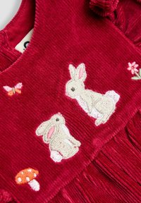Roter Cordstoff mit gestickten Motiven von zwei beigen Kaninchen, einem Schmetterling, einer Blume und einem orangefarbenen Pilz mit weißen Punkten.