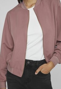 Woman wearing dusty pink zip-up jacket, white t-shirt, and black jeans with one hand in pocket.