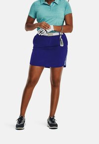 Tenue de golf composée d'un polo bleu sarcelle et d'une jupe violette. La jupe est ajustée et comporte des accents latéraux bleu clair. Des chaussures de golf noires complètent le look.