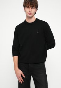 Fekete kötött pulóver bordázott mandzsettával és nyakkivágással, a mellrészen kis logóval. A modell fekete farmert visel, fehér háttér előtt áll.