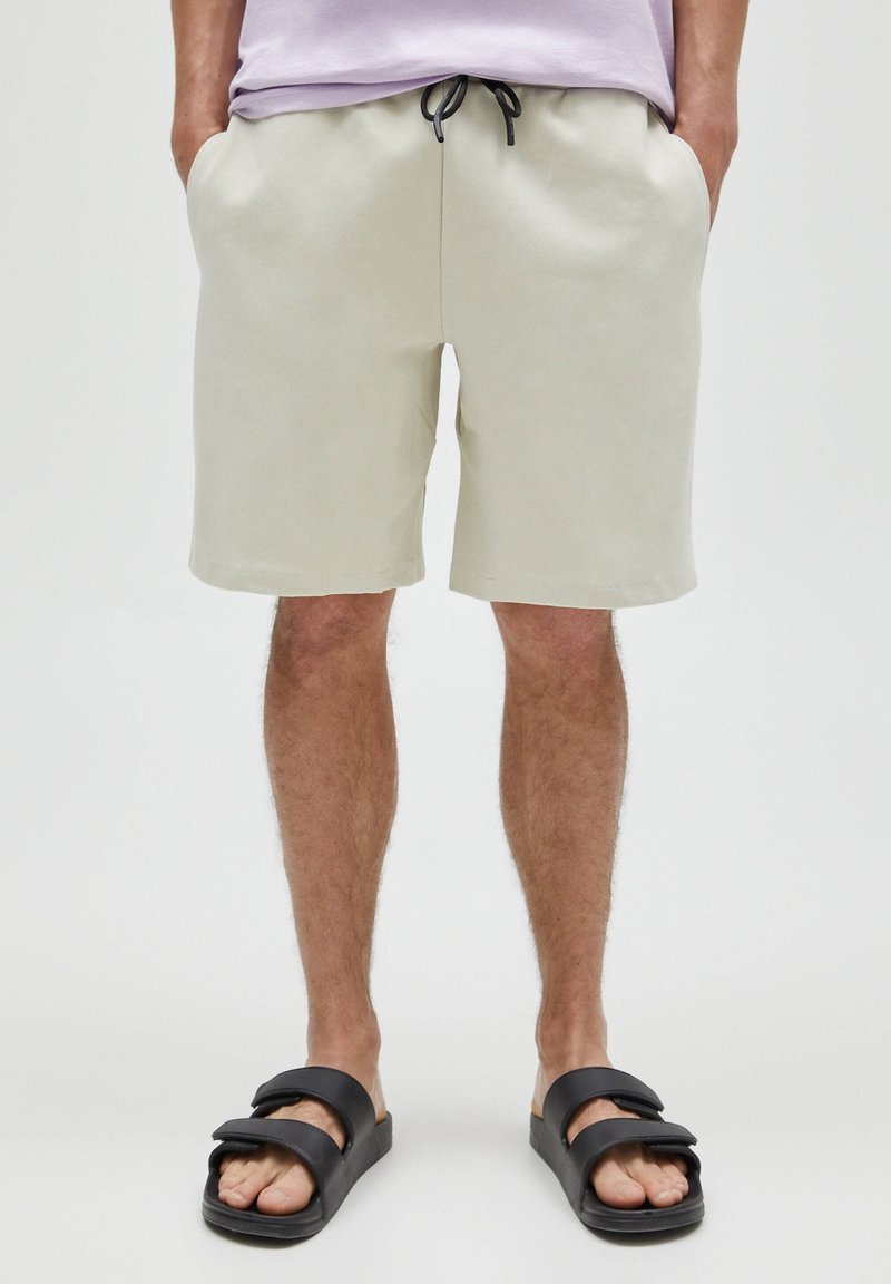 Homme portant un short beige clair à cordon, une chemise lavande et des sandales noires, debout les mains dans les poches contre un fond uni.
