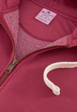 Sudadera de capucha de color burdeos con forro de felpa suave. Presenta un cordón crema y un zipper metálico. Etiqueta de tamaño visible en el interior.