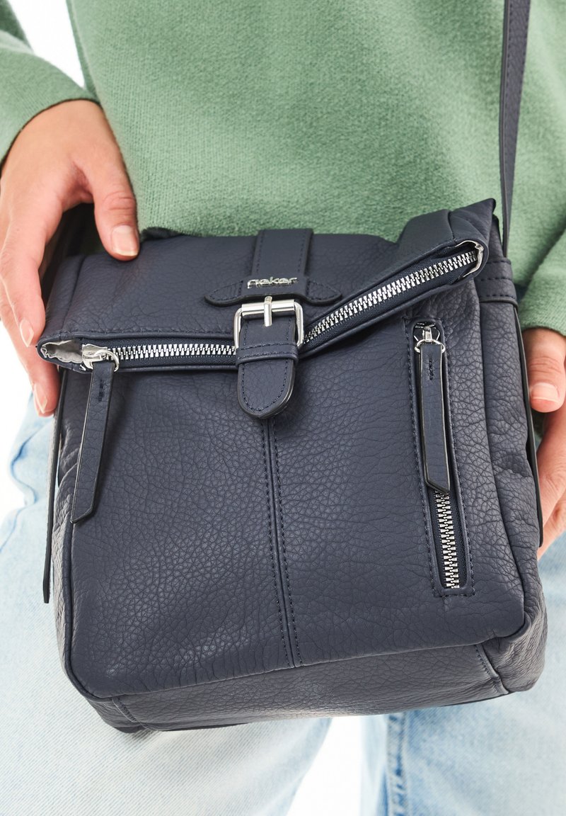 Persona con un suéter verde claro y jeans sostiene un bolso cruzado de cuero texturizado azul marino con detalles de cremallera y hebilla.