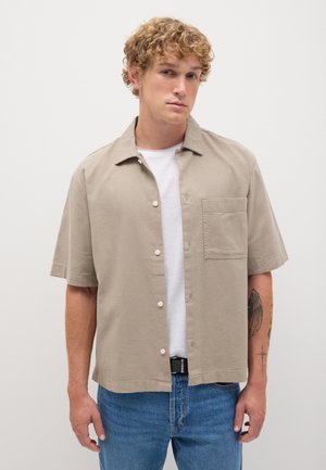 Camicia khaki a maniche corte con bottoni, realizzata in tessuto leggero, dotata di taschino sul petto e vestibilità rilassata, abbinata a jeans blu.