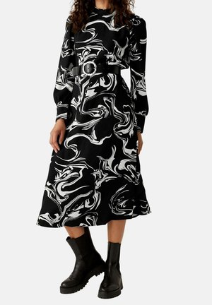 Robe midi noire avec des tourbillons abstraits blancs, manches longues, taille ceinturée, associée à des bottines noires épaisses.