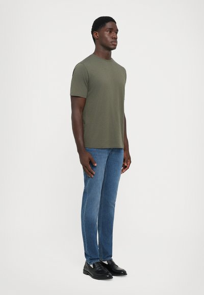Bărbat stând în poziție frontală, purtând un tricou verde măsliniu, blugi albaștri și pantofi de piele negri, pe un fundal alb uniform.