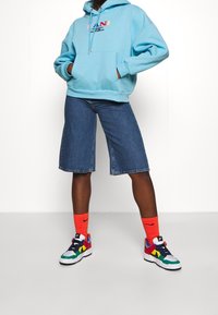 Sudadera azul clara con bolsillo frontal, combinada con pantalones cortos de mezclilla hasta la rodilla. Zapatillas de deporte coloridas y calcetines Nike rojos brillantes completan el look.